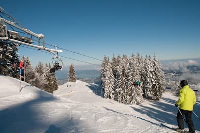A winter scene at Bödele ski resort in Austria featuring a ski lift ascending the snowy mountainside. Nearby a skier embarks on a downhill trek while a charming chalet peeks out from the background.