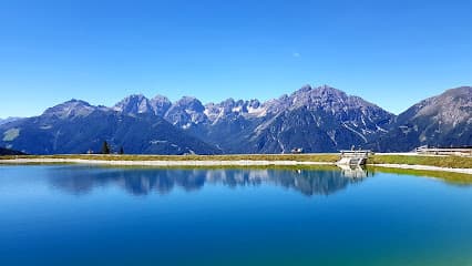 View of a lake reflecting the majestic mountains under a sunny sky at the Serlesbahnen Mieders Bergbahn Bergstation in Innsbruck, Austria, with a cozy chalet nestled nearby and a ski resort in sight.