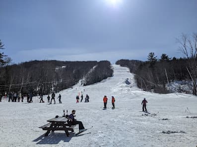 Winter scene at Mt Abram ski resort in Greenwood, Maine, featuring skiers enjoying the slopes. A ski lift is in the background and the landscape is covered in snow.