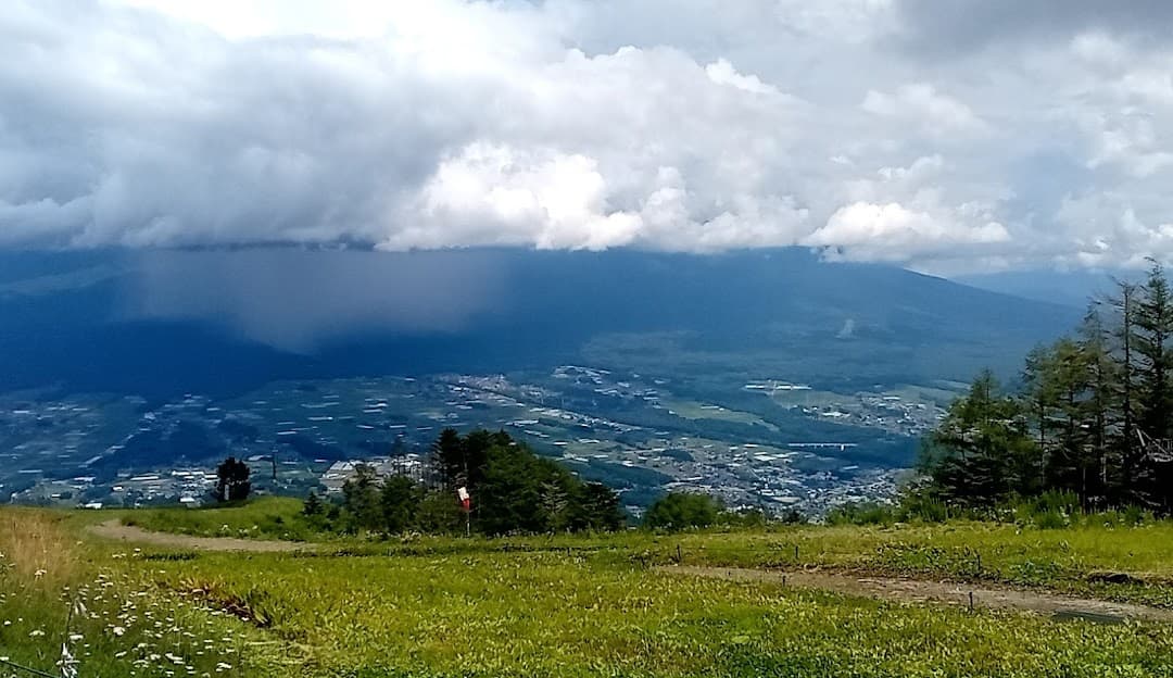 View of the scenic Fujimi Panorama in Nagano, Japan on a sunny day. There's a picturesque mountain with a chalet nestled near the base, while a mountain bike is stationed near. A ski lift adds an adventurous touch to the landscape.