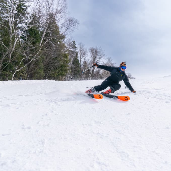 A winter sports scene at Sommet Gabriel in Quebec Canada featuring a skier and a snowboarder on powdery slopes with snowmobile and ski resort vaguely visible.