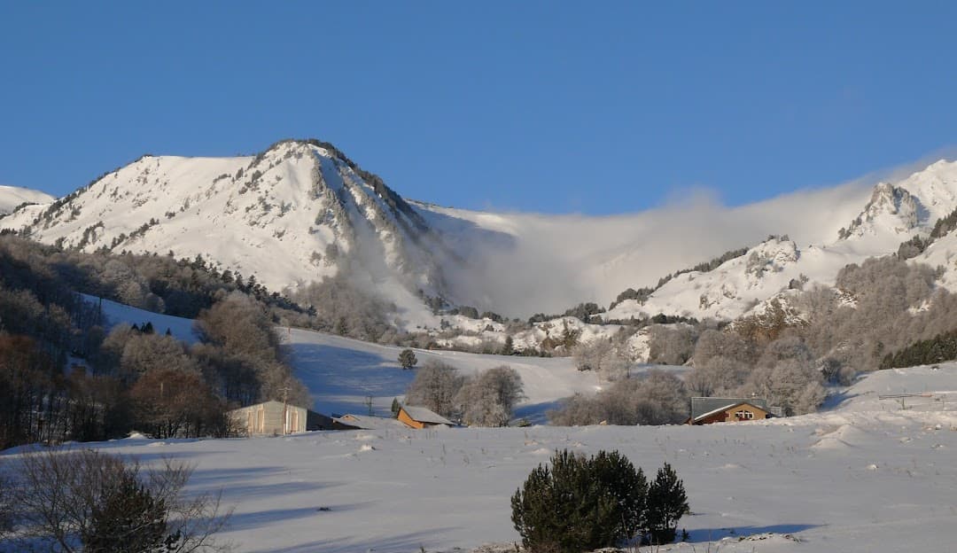 Image of a charming chalet nestled in the snowy winter landscape of Mijanès-Donezan in Ariège, Foix, France. A winter sports centre and mountain are faintly visible in the background.