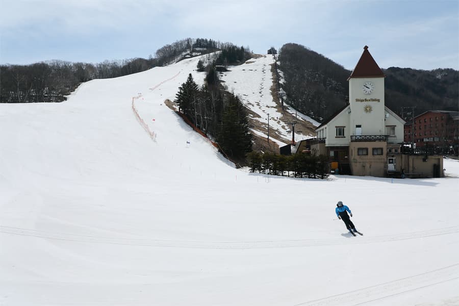 A skier navigating snowy slopes at Hoppo-Bunadaira Ski Area in Yamanochi, Japan. The image features a winter sports scene with a ski resort and lift visible in the background.