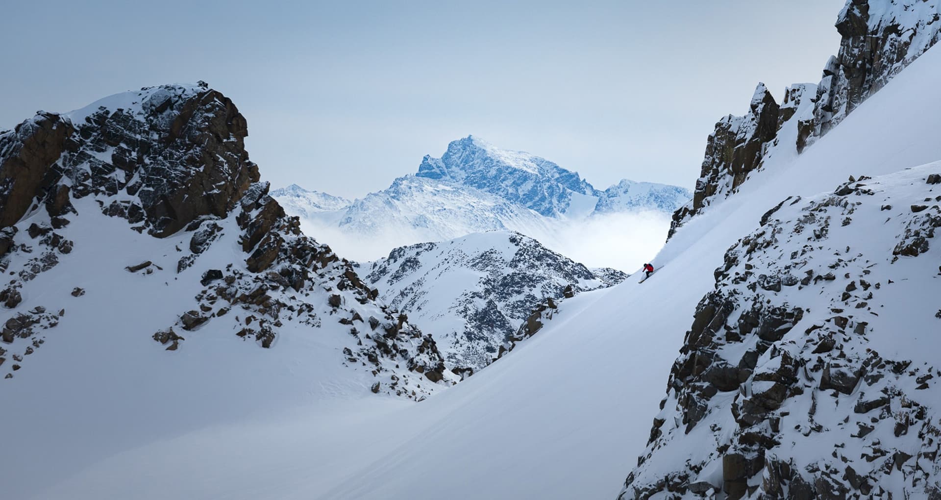 A winter sports scene in Tyax Heliskiing, British Columbia, featuring a skier on a snowy mountain slope near a mountain chalet.