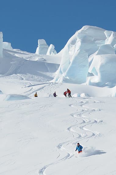 A group of skiers enjoying the winter sports scene at the Tasman Glacier in New Zealand's South Island with a mountain backdrop and a ski resort in the distance.