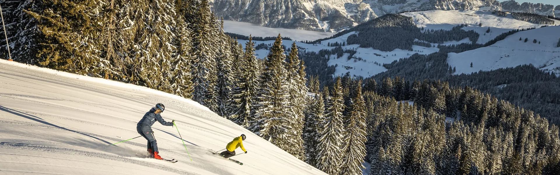 Winter sports scene at SkiWelt-Brixental resort in Tyrol Austria. A skier glides downhill amid a snowy landscape with a snowboarder and a snowmobile in the background.
