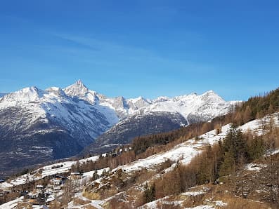 Image depicts a picturesque Swiss ski resort in Valais with a chalet nestled against a snow-covered mountain backdrop. Winter sports enthusiasts can be seen partaking in the outstanding facilities available.